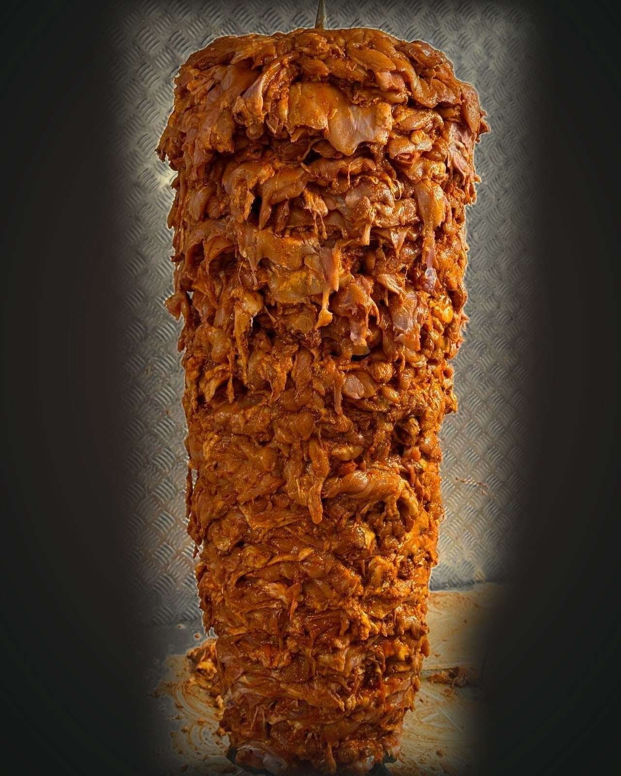 Whole chicken doner, raw and marinated, on its skewer against a steel wall